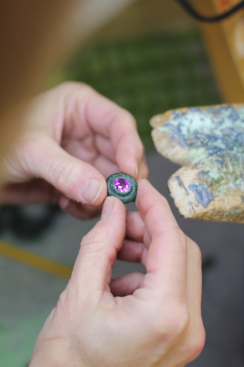 Hands holding a green wax jewelry model with a purple gemstone at the studio workbench